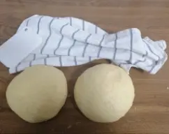 Step <p>Con la espátula, baje la masa hacia el fondo del vaso y vuelva a amasar durante 1 min sin poner el cubilete. Retire la masa del vaso (estará blanda) y póngala sobre la superficie de trabajo. Dele forma de bola y divida la masa en 2 partes, dejarlas reposar durante unos 5 minutos tapadas con un trapo de cocina, volver a formar de nuevo la bola y dejarla reposar otros 5 minutos. Forrar dos bandejas de horno con papel de hornear.</p> illustration